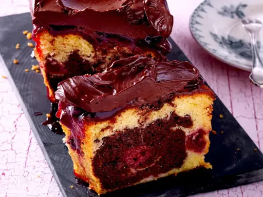 Zwei Stücke saftiger Marmorkuchen mit Kirschen und Schokoladenglasur auf einem dunklen Brett.