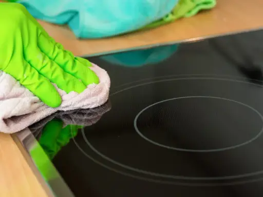 Grüne Handschuhe wischen mit einem Tuch über das Induktionskochfeld, um es zu reinigen.