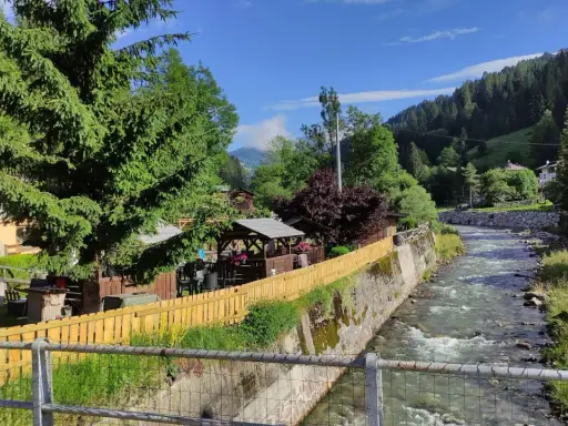 Un ruscello scorre a Santo Stefano di Cadore, fiancheggiato da case, alberi e un'area con gazebo e tavoli all'aperto.