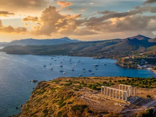 Rovine del Tempio di Poseidone a Capo Sounion, con barche a vela che navigano nel mare blu sotto un cielo dorato al tramonto.