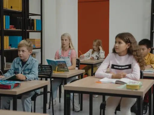 Dzieci siedzą w klasie, skupione na lekcji. Chłopiec w dżinsowej kurtce, dziewczynka z książką po angielsku, a z tyłu inne dzieci. Wszyscy mieli nadzieję, że lekcja szybko się skończy.