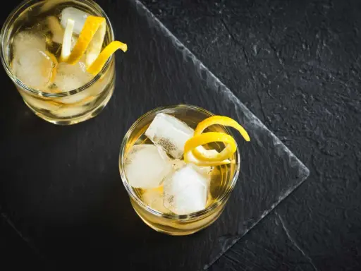 Zwei erfrischende Whiskey Cocktails mit Eis und Zitronenzesten auf dunklem Stein.