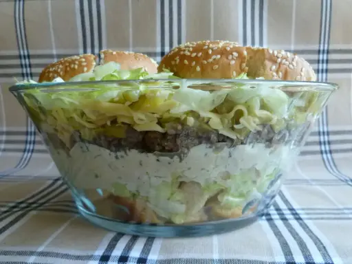 Warstwowa sałatka big mac w szklanej misie, z bułkami na wierzchu.