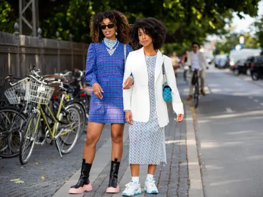 Two women showcase their spring chic dress code. One wears a blue plaid dress and chunky boots, the other a white blazer over a patterned dress and sneakers.