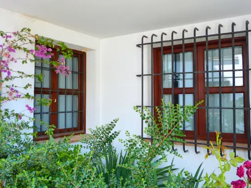 Dos ventanas con barrotillo de hierro y marcos de madera, rodeadas de vegetación y flores moradas.