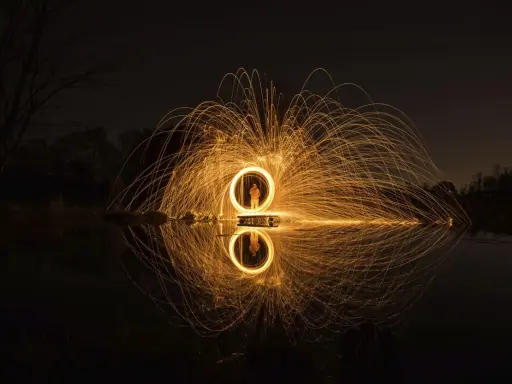 Un cerchio di scintille dorate, un uomo al centro, riflette sull'acqua. Un'opera d'arte che sembra **disegnare con la luce** nel buio della notte.