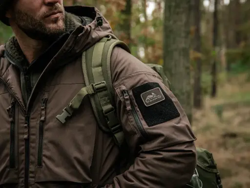 Mężczyzna w brązowej kurtce z naszywką Helikon-Tex, symbol polskie marki sportowe, w lesie.