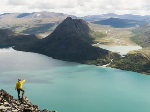 En person i gul jacka pekar ut över en turkos sjö och berglandskap under en Jotunheimen vandring.