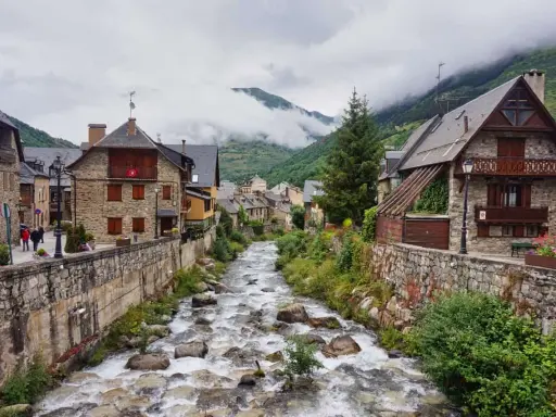Ein rauschender Fluss schlängelt sich durch das malerische Val d'Aran, gesäumt von traditionellen Steinhäusern und grünen Bergen.