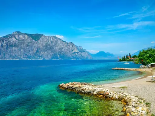 Ein idyllischer Gardasee Strand mit kristallklarem Wasser, felsigen Ufern und majestätischen Bergen im Hintergrund.