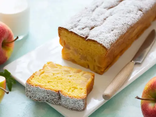 Un delizioso plumcake alle mele, cosparso di zucchero a velo, con una fetta già tagliata. Mele rosse e un bicchiere di latte completano la scena.