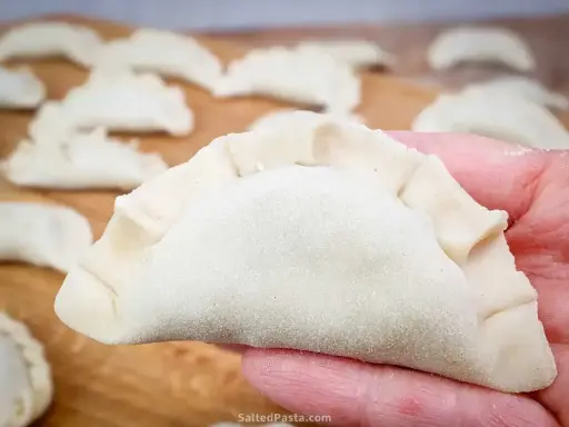 Dłoń trzyma surowe pierogi z serem, gotowe do gotowania. Na drewnianej desce widać więcej pierogów.
