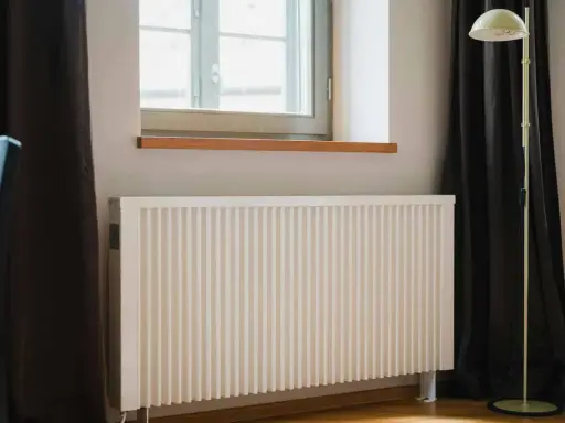 Biały grzejnik pod oknem, obok ciemnej zasłony i lampy stojącej. Dowiedz się, jak ustawić grzejniki przy ogrzewaniu gazowym dla optymalnego komfortu.