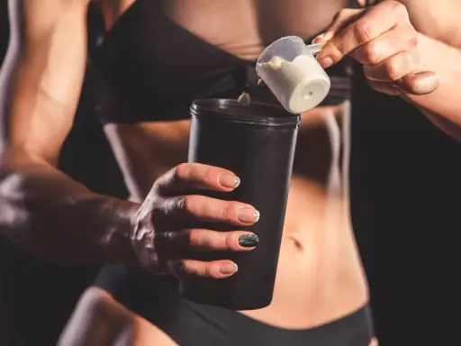 Kobieta z umięśnionym brzuchem dodaje proszek do shakera. Czy shake proteinowy jest zdrowy? To zależy od składu.