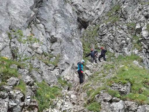 Wspinaczka w skalistym terenie, przypominającym wejście do jaskini raptawickiej. Trzy osoby w kaskach pokonują trudną trasę.