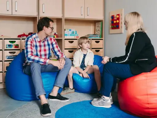 Ojciec, syn i terapeuta dla dziecka rozmawiają w przytulnym pokoju. Na ścianie wisi tablica sensoryczna.