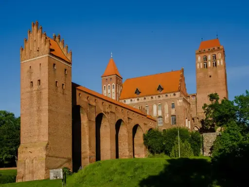Potężny zamek w Kwidzynie z cegły, z charakterystycznymi wieżami i arkadowym murem obronnym, lśni w słońcu na tle błękitnego nieba.
