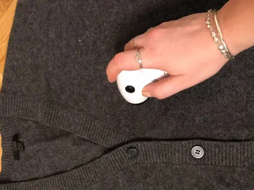 En hand med smycken använder en noppmaskin på en mörkgrå kofta. Materialet känns mjukt, kanske rayon material svenska.