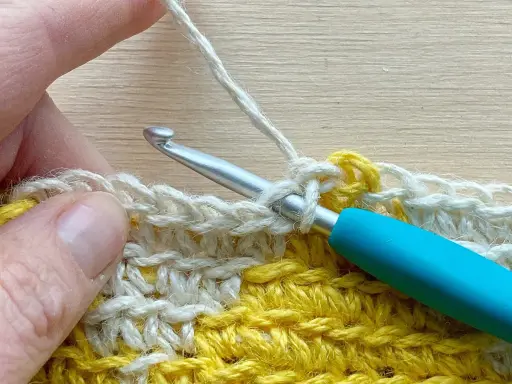 Mano tejiendo con ganchillo, creando un punto bajo con hilo blanco sobre una tela amarilla y blanca.
