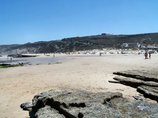 Sonniger Strand bei Monte Clerigo mit Felsen und Menschen, die sich im Wasser und am Ufer aufhalten.