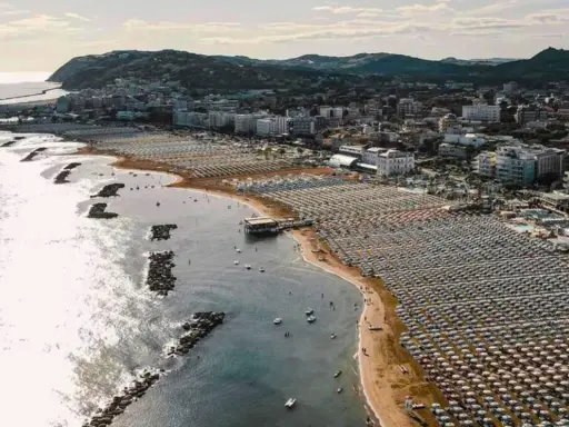 Vista aerea della cattolica spiaggia, con file ordinate di ombrelloni e lettini che si estendono lungo la costa, mentre il mare luccica sotto il sole.
