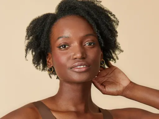 Une femme à la peau foncée, aux cheveux afro volumineux et bouclés, porte des boucles d'oreilles dorées.