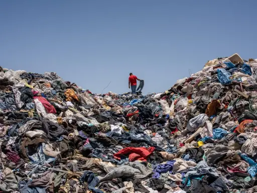 Un uomo in maglia rossa cammina su una montagna di vestiti scartati, simbolo dell'inquinamento tessile.