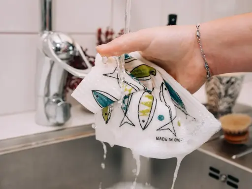 En hand sköljer en disktrasa med fiskmönster under rinnande vatten. Trasan är gjord av garn till disktrasa och har texten "Made in Sweden".