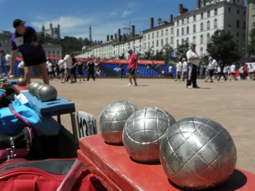 Boules de jeu en gros plan, prêtes pour une partie de boule lyonnaise. Des joueurs s'affrontent en arrière-plan.