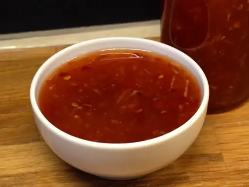En skål med glansig, röd sweet chili sås med små bitar av chili och vitlök. En flaska med samma sås syns i bakgrunden.