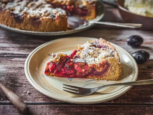 Ein saftiger Zwetschgenkuchen mit Dinkelmehl vom Blech, bestäubt mit Puderzucker. Ein Stück auf einem Teller mit einer Gabel.