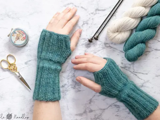 Manos con guantes tejidos a mano, listos para un nuevo patron. Lana, agujas y tijeras decorativas completan la escena.
