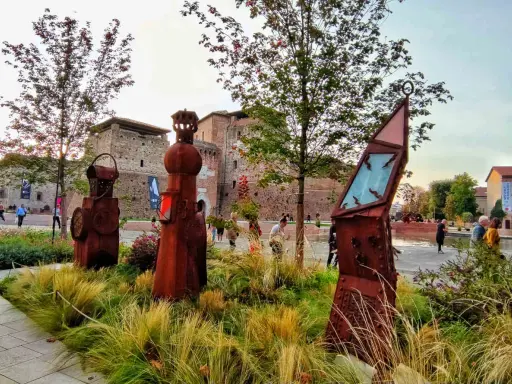 Sculture arrugginite e vegetazione rigogliosa nel Parco Fellini a Rimini, con il castello Sismondo sullo sfondo.