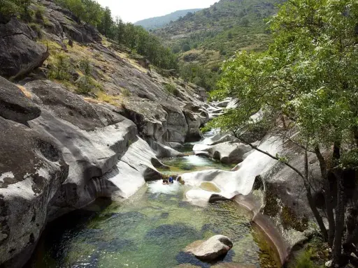 Un río de aguas cristalinas fluye entre rocas erosionadas, formando piscinas naturales. Dos personas disfrutan del baño en uno de los pilones, rodeados de vegetación y montañas.