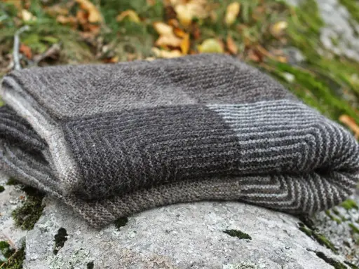 En hopvikt, handstickad filt i gråa nyanser ligger på en mossig sten. Perfekt för mysiga höstkvällar.