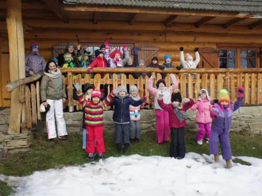 Wesoła gromadka dzieci pozdrawia z bacówki u harnasia. Wszyscy ubrani w zimowe stroje, radośnie machają rękami na tle drewnianego domu.