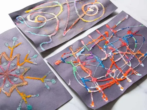 Kinder malen auf schwarzem Papier mit bunten Salzkristallen. Ein Bild zeigt eine Schneeflocke, die anderen abstrakte Muster.