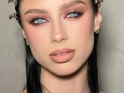 Ojos ahumados en tonos rosados y marrones, realzando unos ojos azules penetrantes. Labios nude y cabello oscuro completan el look.
