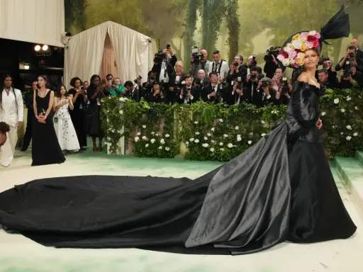 Una mujer con un vestido negro y un tocado floral posa para las cámaras en la alfombra verde, mientras un hombre ajusta la cola de su vestido, todo bajo el tema de la Met Gala.