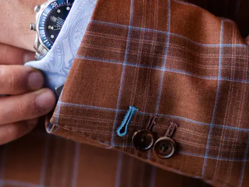 A man adjusts his watch on his wrist, revealing the detailed pick stitching on the cuff of his brown plaid suit jacket.