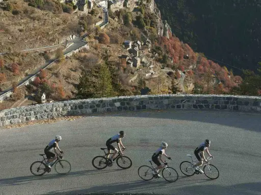 Ciclisti affrontano la salita del Col de la Couillole, con un villaggio arroccato sulla montagna autunnale.