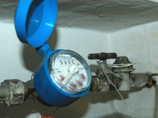 Contatore acqua blu con coperchio aperto, il quadrante è illeggibile per condensa. Tubi e rubinetto arrugginiti.