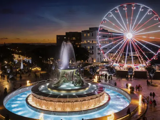 Una piazza illuminata a festa a Malta a Natale, con una fontana scintillante e una ruota panoramica che domina lo skyline al tramonto.
