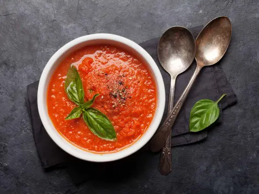 Schüssel mit selbstgemachter Tomatensauce, garniert mit Basilikum und Pfeffer, daneben zwei Löffel auf dunklem Hintergrund.