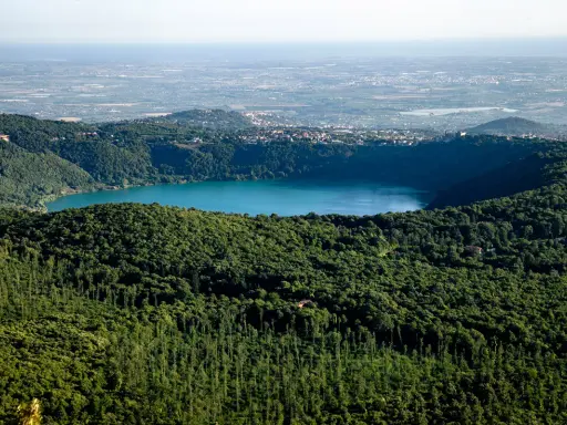 Vista panoramica del lago di Albano, circondato da rigogliosa vegetazione, con il borgo di Castel Gandolfo e Rocca di Papa in lontananza.