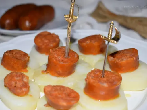 Chorizo a la sidra en rodajas, servido sobre patatas y adornado con espadas doradas.