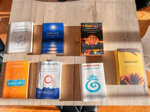Una collezione di **libri** di Gianluca Gotto disposti su un tavolo di legno. Tra i titoli: "Le coordinate della felicità", "Come una notte a Bali", "La Pura Vida".