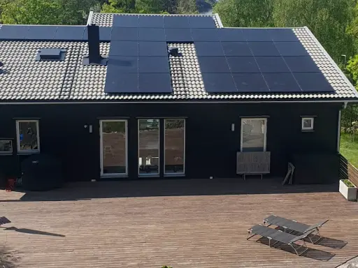 Ett mörkt hus med en liten solcellsanläggning på taket. En stor altan med solstolar leder ut mot en grönskande trädgård.