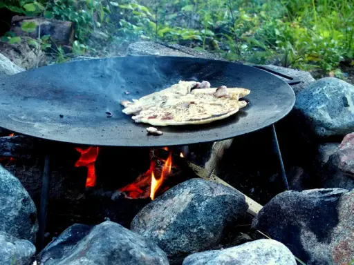 Matlagning över öppen eld på ett muurikka ställning. Pannkakor steks i en stor stekpanna över glödande kol och ved.