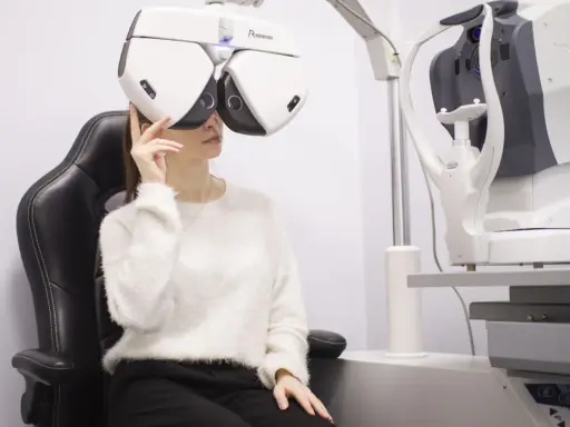 Kobieta w białym swetrze siedzi na czarnym fotelu, badana przez optometrystę na nowoczesnym sprzęcie.